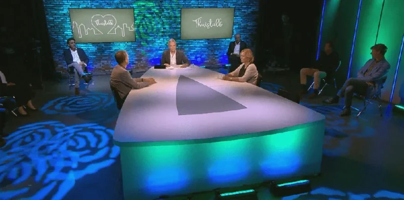 Studiodecor met een ronde verlichte tafel en gasten in gesprek, in groen uitgelicht decor.