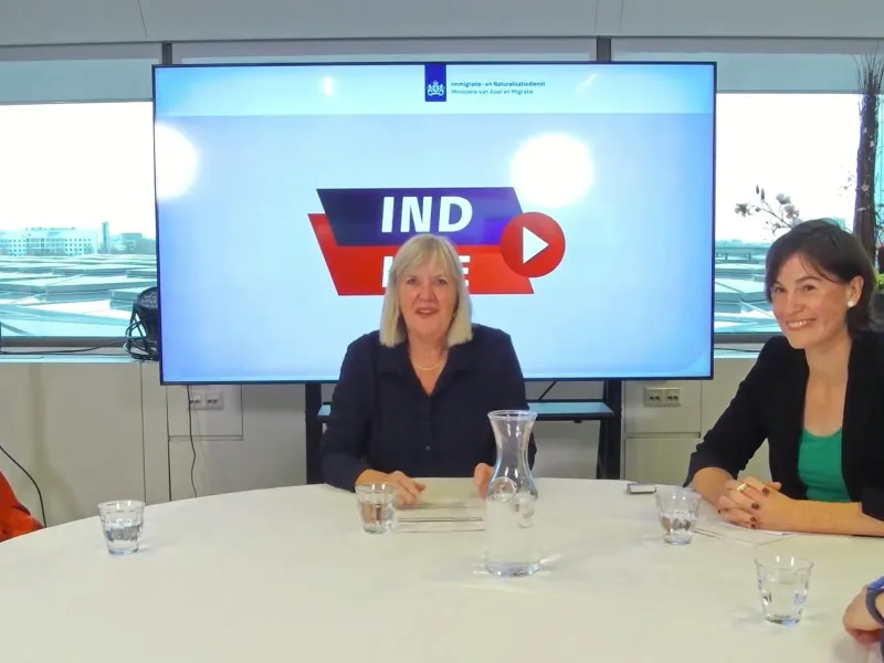 Paneelgesprek aan een ronde tafel tijdens een livestream over actualiteit en beleid, met de presentatie op een groot scherm.