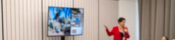 Spreker voor een scherm in een zaal met een groep toehoorders, terwijl de sessie live wordt uitgezonden.