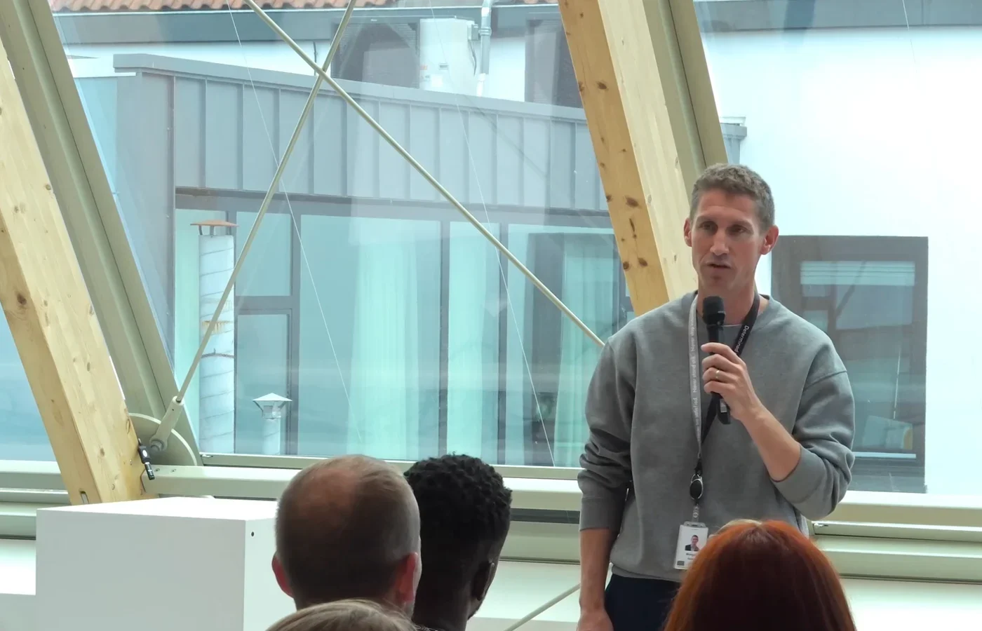 Spreker met microfoon richt zich tot collega's in een lichte zaal met houten balken, tijdens een marketingupdate.