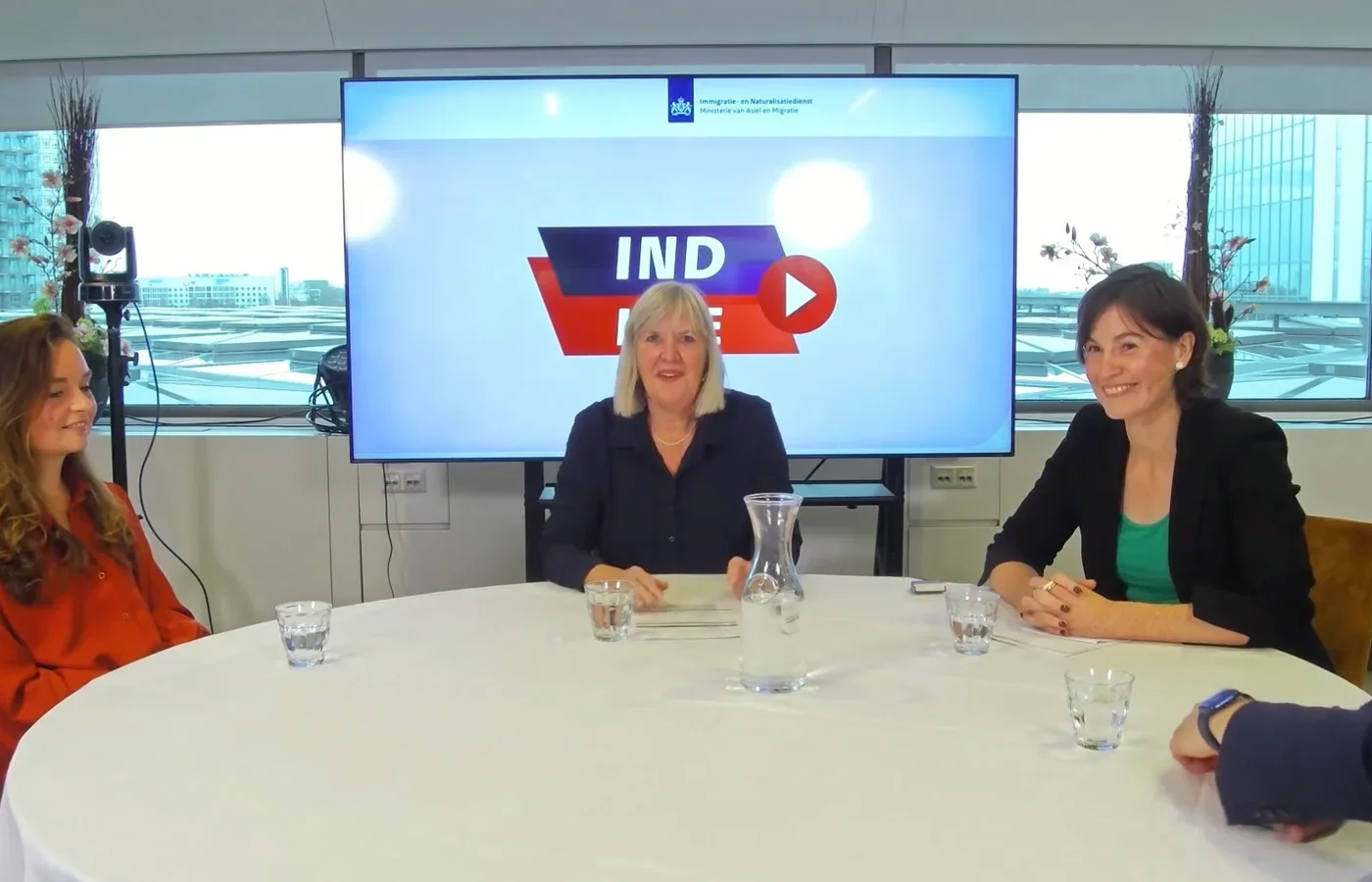 Drie gespreksdeelnemers aan een ronde tafel met het IND-scherm achter zich, tijdens een interne uitzending.