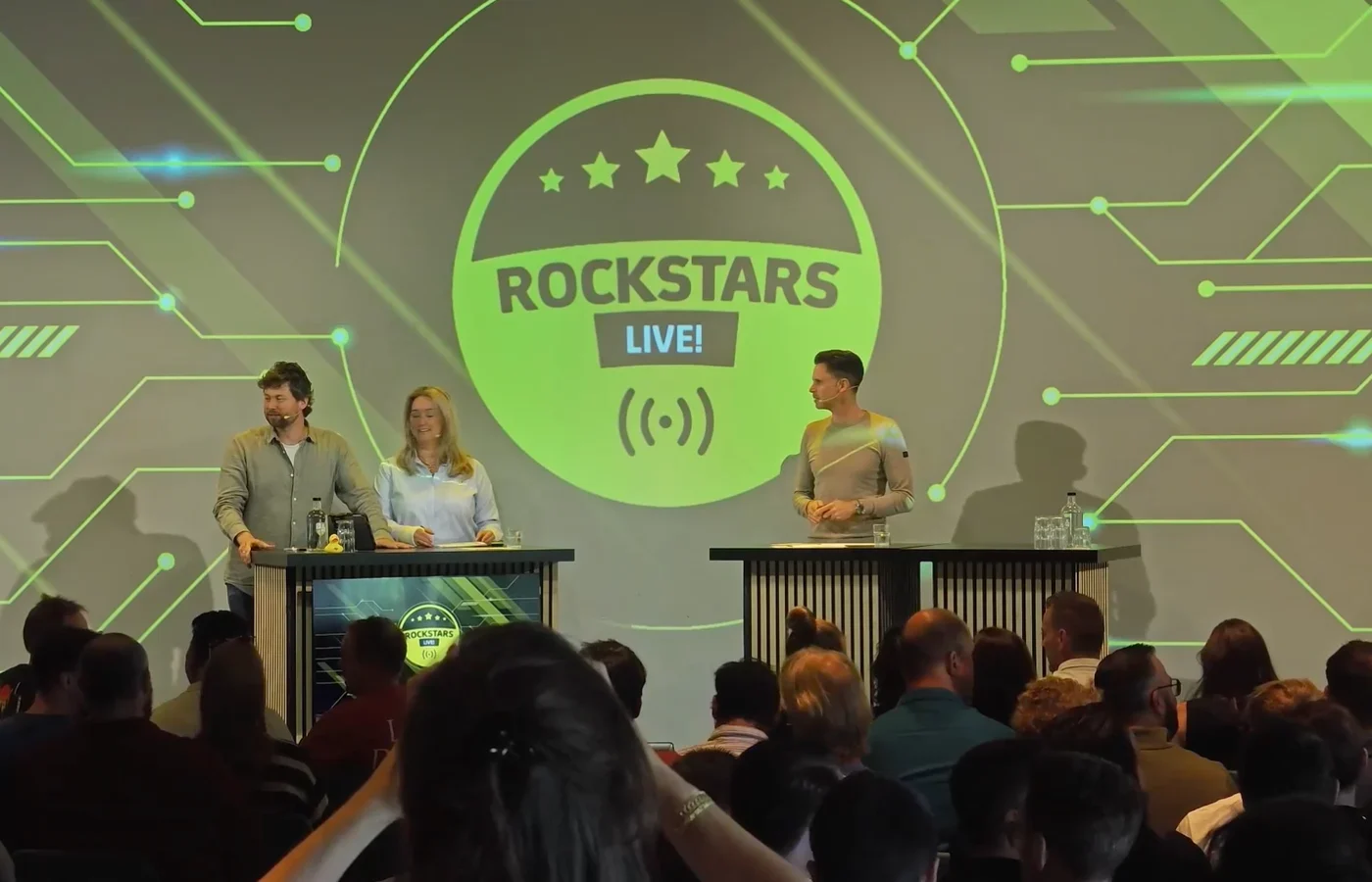 Twee presentatoren op het verlichte podium van Rockstars Live, met het publiek in de zaal ervoor.