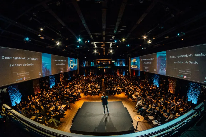 Volle zaal met een spreker op een rond podium en panoramische schermen rondom het publiek.