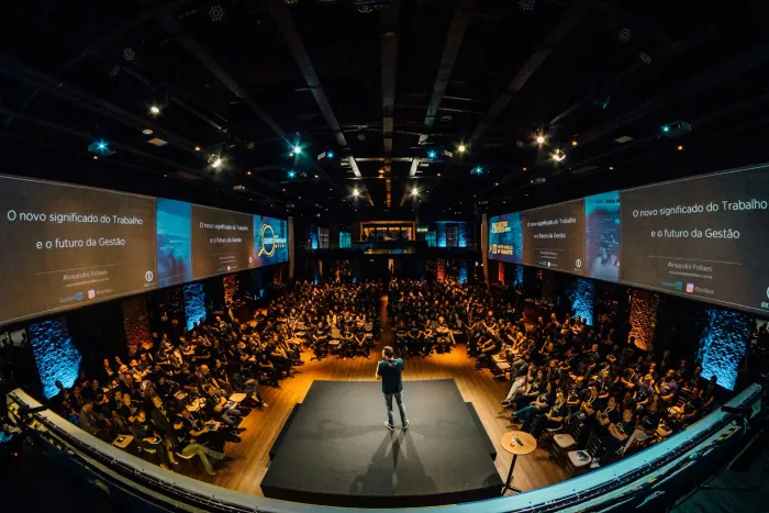 Volle zaal met een spreker op een rond podium en panoramische schermen.