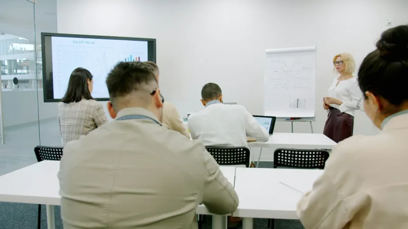 Deelnemers aan tafels volgen een presentatie met grafieken op een scherm in een lichte vergaderruimte.