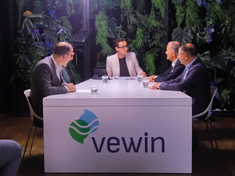 Gespreksleider en drie gasten aan een studiotafel voor een groene plantenwand, tijdens de opname van een talkshow voor Vewin.