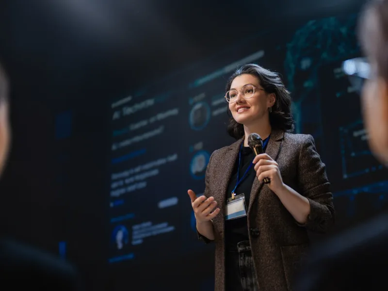 Spreker met headsetmicrofoon voor een groot donker scherm tijdens een keynote.