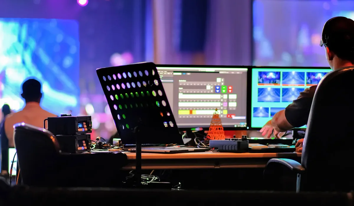 Regieplek met multiviewmonitoren en een lichttafel, terwijl het podium op de achtergrond wordt uitgelicht.