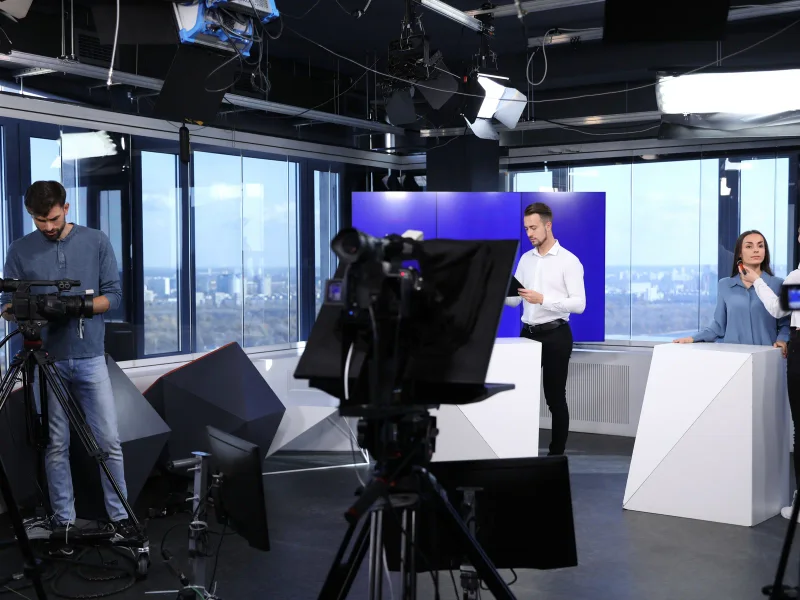 Cameraploeg bouwt een studioset op met camera's op statief, studiolampen en een teleprompter rond twee presentatoren.