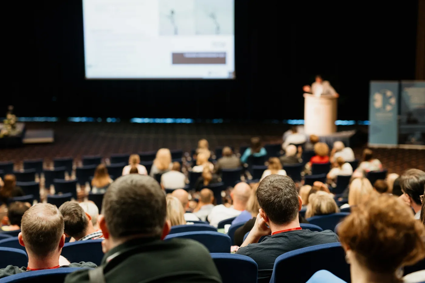 Publiek in een congreszaal kijkt naar een spreker op het podium met een groot presentatiescherm erboven.