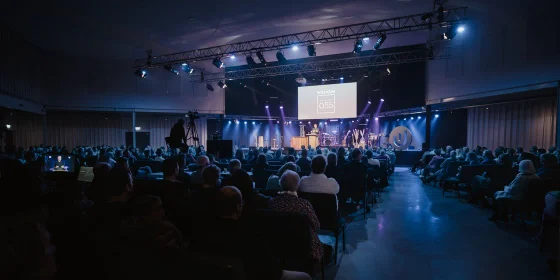 Volle zaal kijkt naar een verlicht podium tijdens een programma.
