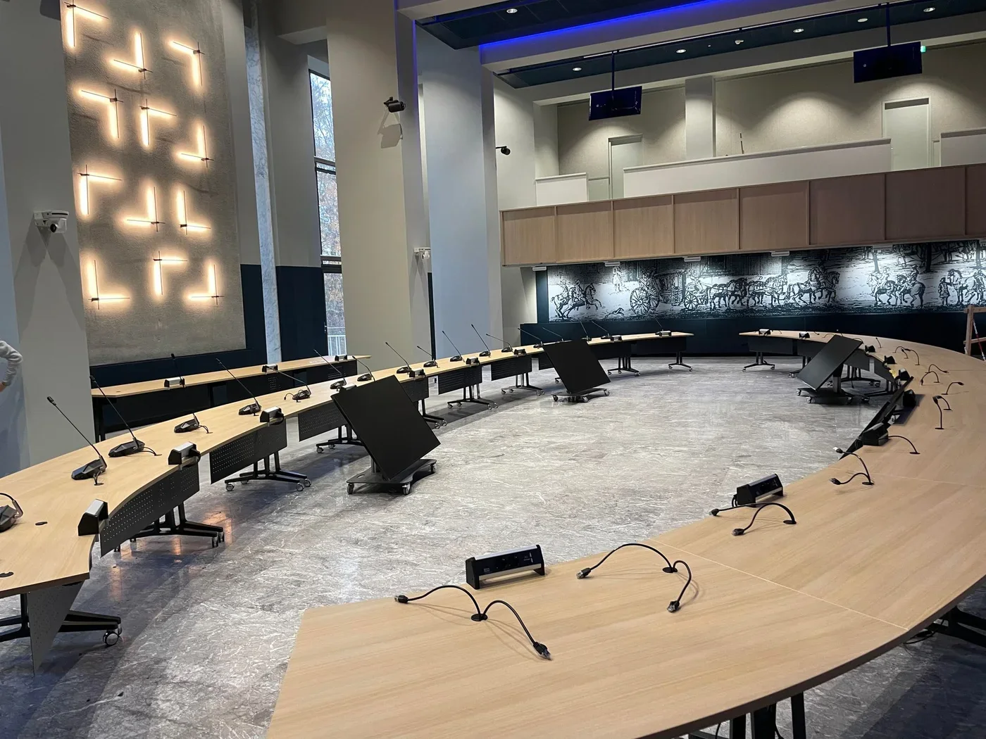Lege raadszaal met een hoefijzeropstelling van tafels en microfoons, klaar voor de uitzending van een vergadering.