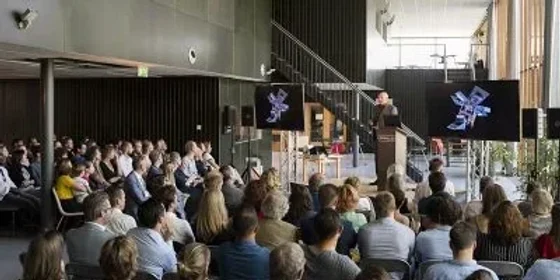 Publiek in een zaal kijkt naar een spreker bij een presentatiescherm.