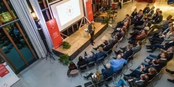 Zaal van bovenaf met publiek, podium en een groot presentatiescherm.