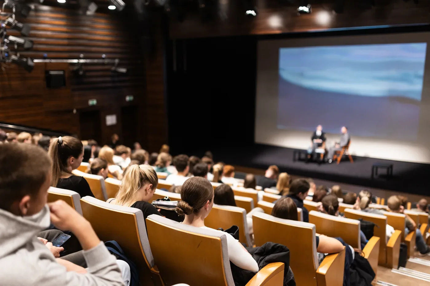 Volle collegezaal waar studenten kijken naar een gesprek op het podium met een groot projectiescherm.