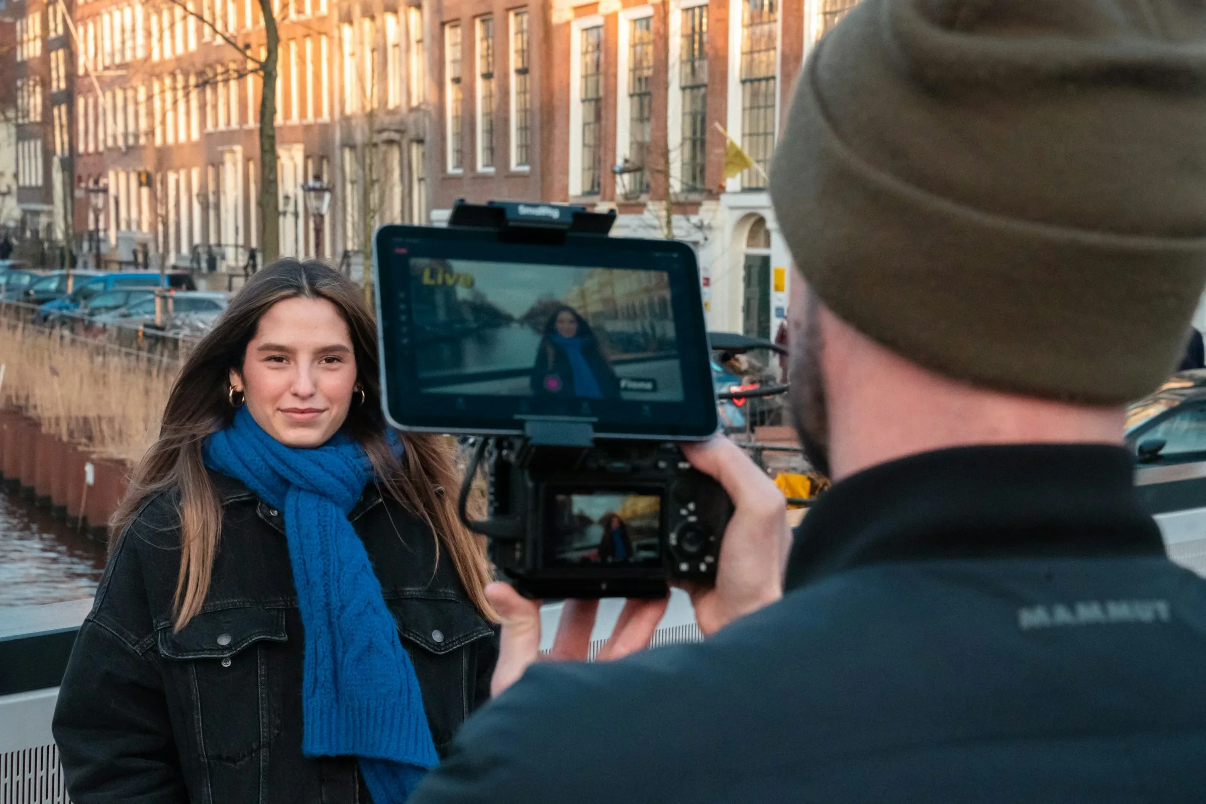 Cameraman filmt een presentatrice aan een Amsterdamse gracht, met de live opname op de monitor van de camera.