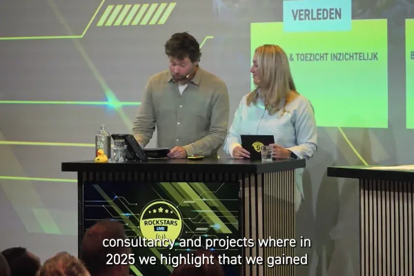 Studio-uitzending met twee presentatoren achter een desk en ondertiteling onder in beeld.