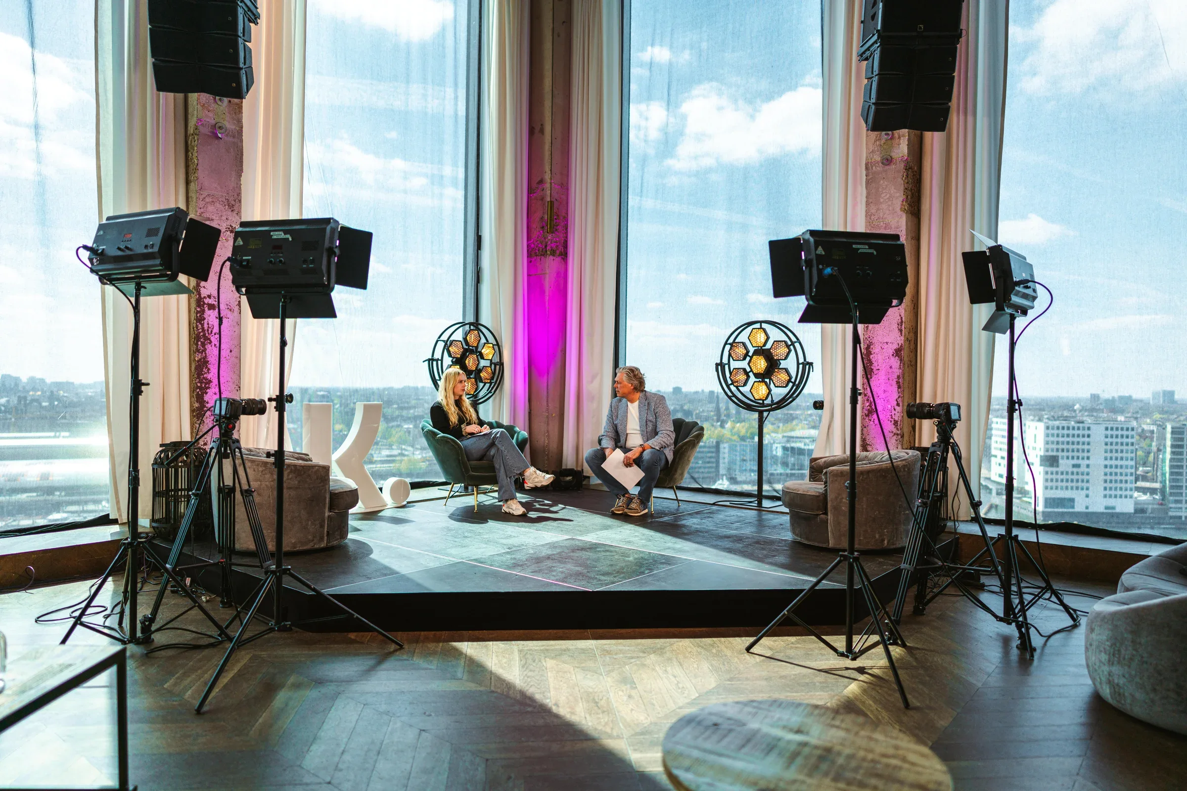 Studioset met camera's, studiolampen en een zithoek voor grote ramen met uitzicht over de stad.