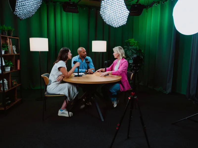 Vier mensen in gesprek rond een ronde tafel in een groen uitgelichte studio, met softboxen en een camera op statief.