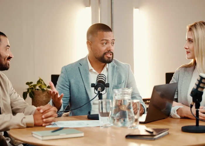 Vier gesprekspartners rond een tafel met microfoons en een laptop, in een lichte kantoorruimte waar de podcaststudio is opgebouwd.