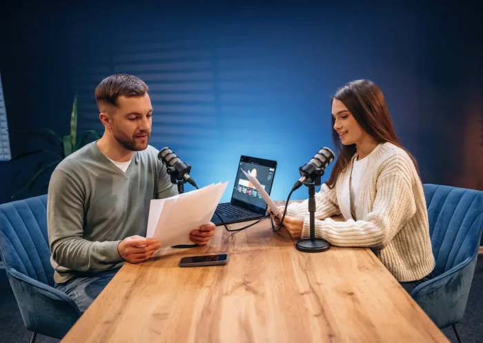 Twee presentatoren aan een houten tafel met microfoons en een laptop, in een blauw uitgelichte podcaststudio.
