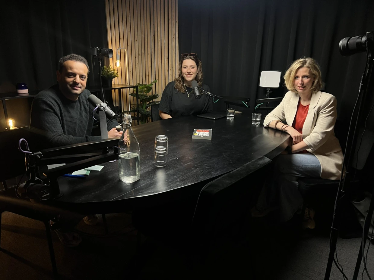 Podcastopname in de studio: drie gasten aan een tafel met microfoons op statief, professionele belichting en een camera opgesteld voor een videopodcast.