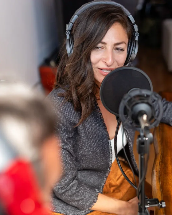 Gast met koptelefoon op vertelt haar verhaal voor de microfoon met plopkap, terwijl haar gesprekspartner op de voorgrond meeluistert.