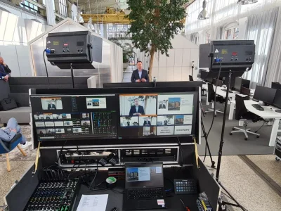 Regieopstelling met multiviewmonitoren en een geluidstafel in een atrium, met studiolampen en een spreker op de achtergrond.