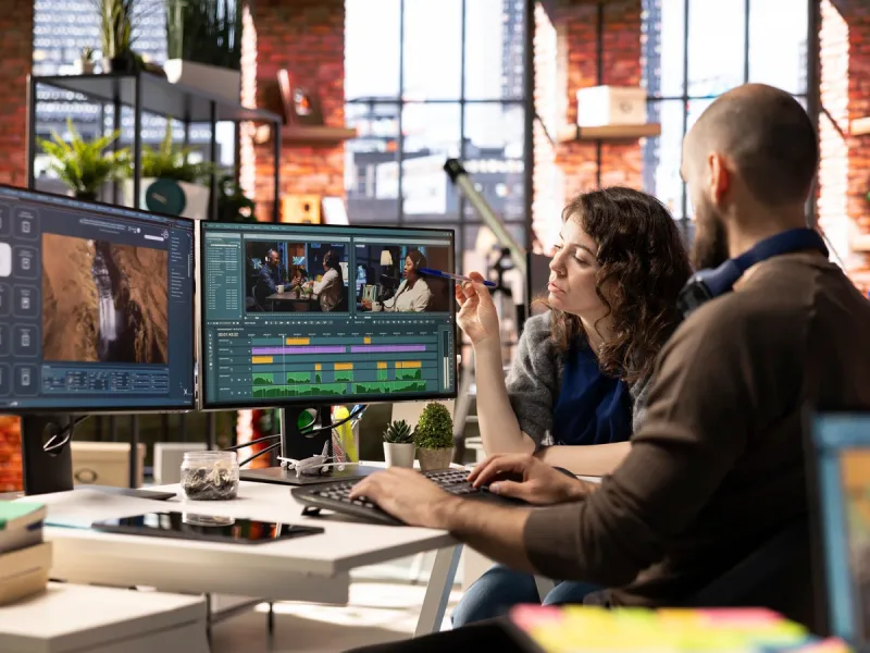 Editor bewerkt de opname op een montagescherm in een kantoor, met collega's op de achtergrond.