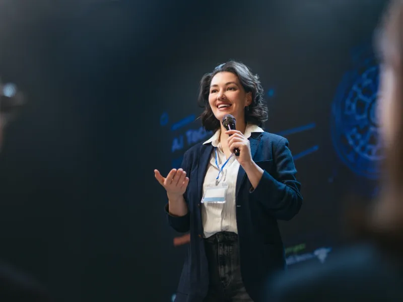Spreker met microfoon op een podium in een verduisterde zaal, terwijl het publiek meeluistert.
