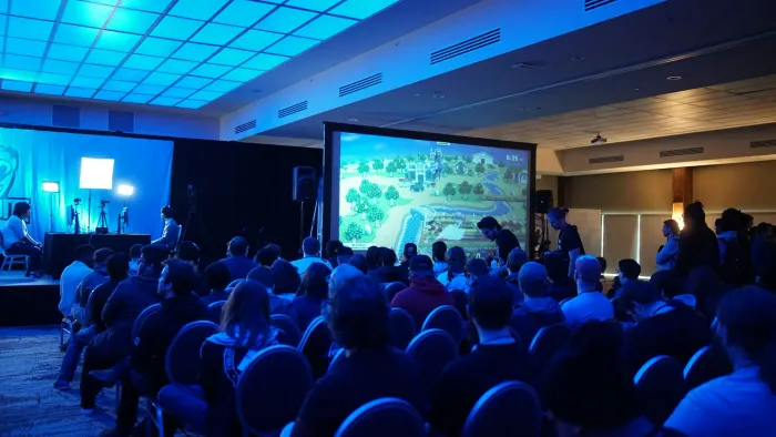 Publiek in een blauw uitgelichte zaal kijkt naar een groot presentatiescherm.