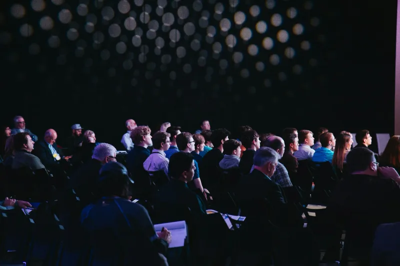 Publiek in een verduisterde zaal kijkt naar het podium tijdens een internationaal event.