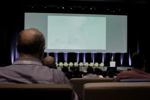 Publiek in de zaal kijkt naar het podium met sprekers en een groot scherm.