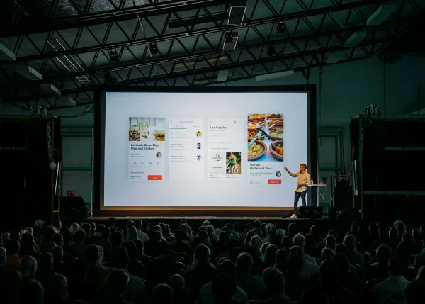 Spreker op een podium voor een groot scherm in een zaal met honderden bezoekers.
