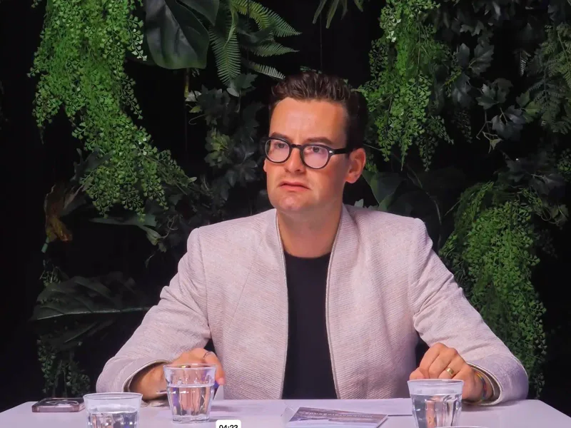 Presentator aan tafel in een livestream studio met groene plantenwand, tijdens de opname van een talkshow.