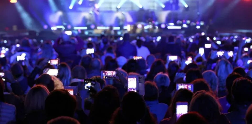 Publiek met telefoons in de lucht in een blauw uitgelichte zaal tijdens een evenement.