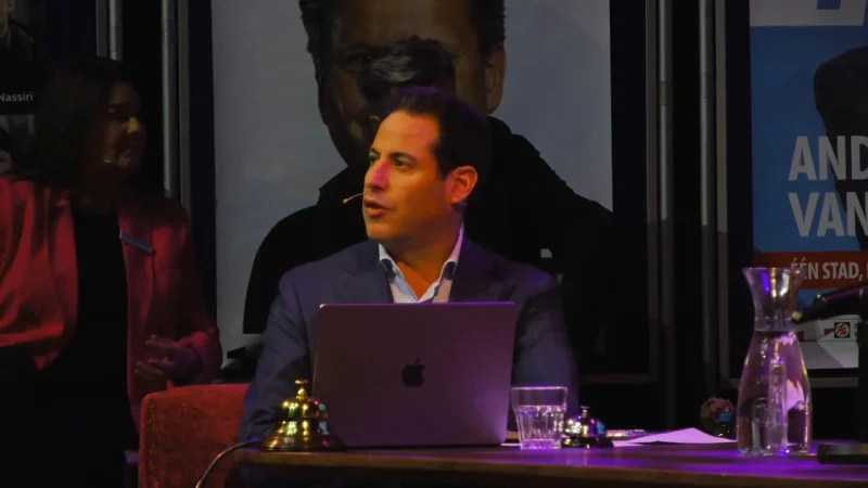 Spreker achter een laptop aan tafel tijdens een bijeenkomst, met een verlicht portret op de achtergrond.