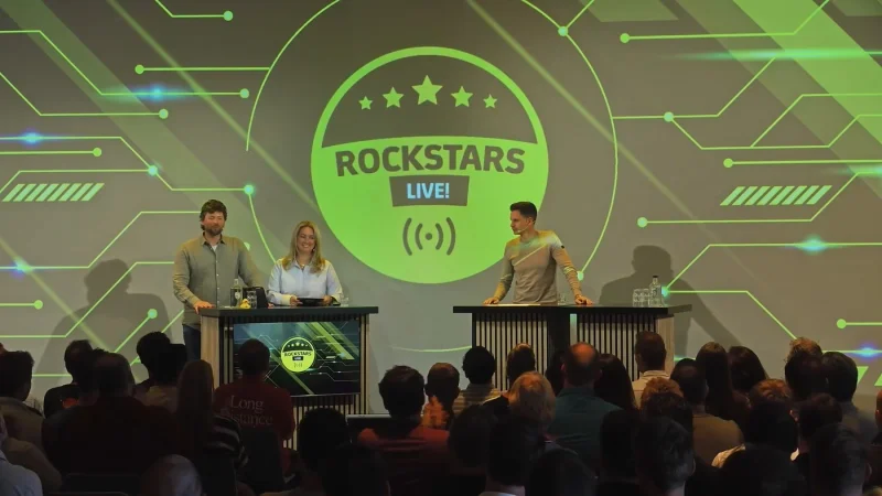 Twee presentatoren en een gast op het podium van Rockstars Live, met het publiek in de zaal ervoor.