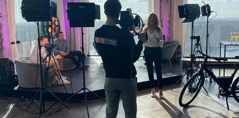 Crew filmt een gesprek bij het raam, met camera's, statieven en studiolampen.