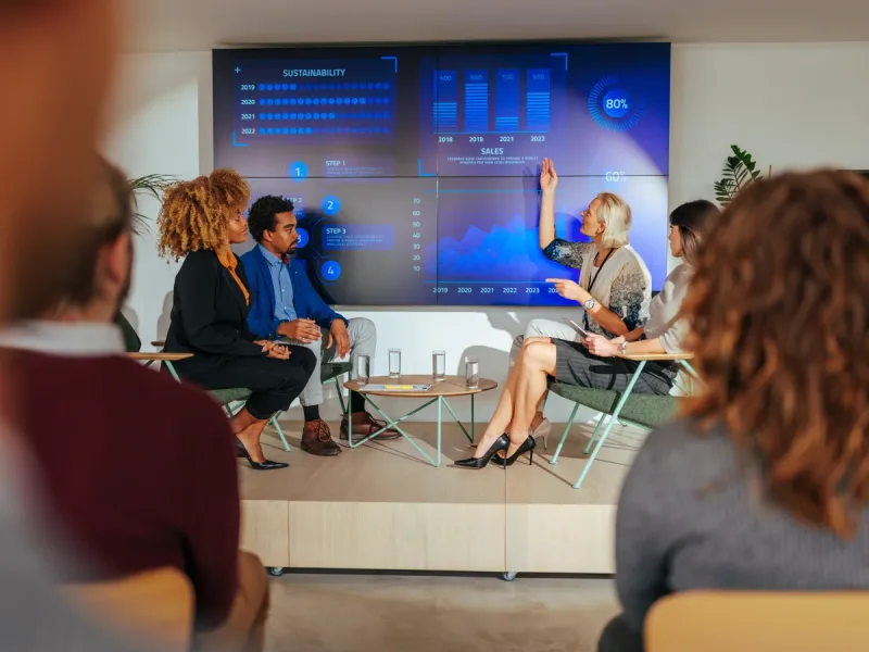 Panel van sprekers op een podium voor een groot dashboardscherm, terwijl collega’s vanaf de zaal meekijken tijdens een interne uitzending.
