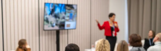Groep deelnemers volgt in een zaal een spreker die het webinar vanaf locatie presenteert.