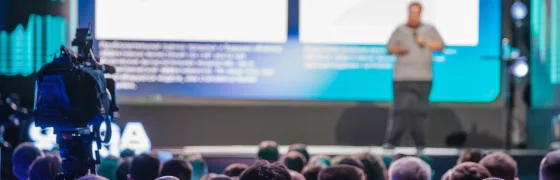 Volle congreszaal met grote projectieschermen en een camera vooraan, waar het webinar internationaal wordt gevolgd.