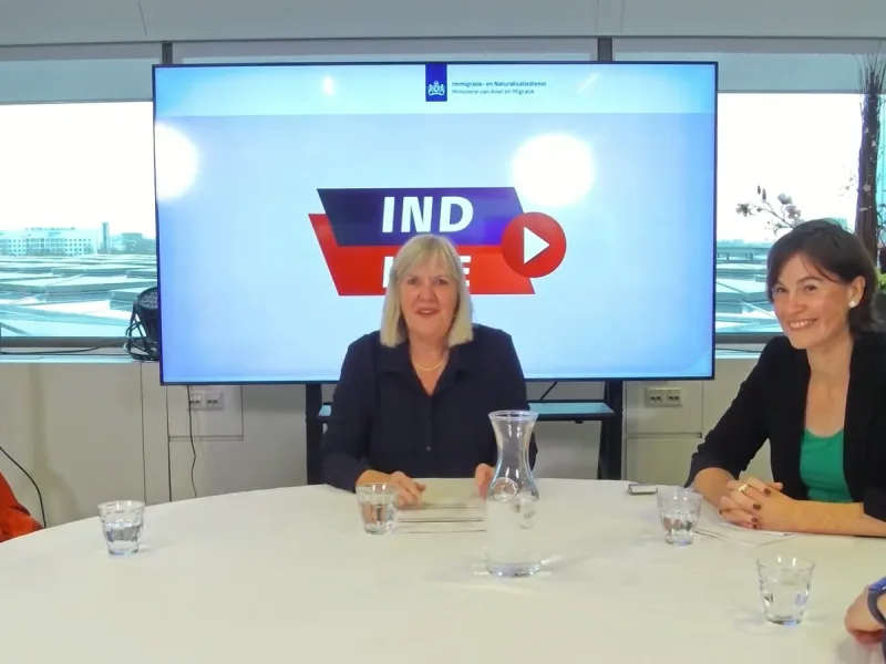 Twee gespreksdeelnemers aan een ronde tafel met een groot scherm van een overheidsdienst achter zich.