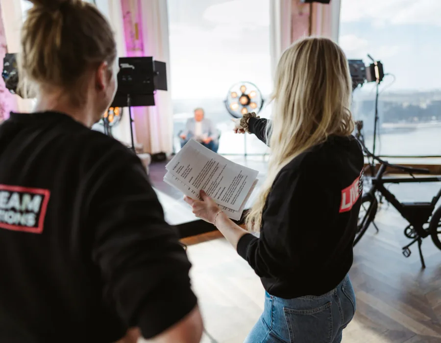 Twee crewleden van Livestream Solutions overleggen met het draaiboek in de hand op de set, met studiolampen en de spreker op de achtergrond.